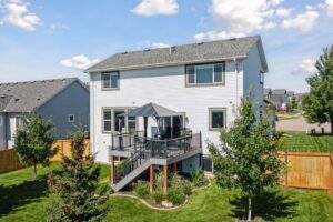 Two-story white house with a gray roof at 15668 Fairchild Way. Enjoy the fenced backyard with an elevated deck, patio furniture, and umbrella. Stairs lead to the green lawn and trees, all under a blue sky. See PHOTOS for more details!.