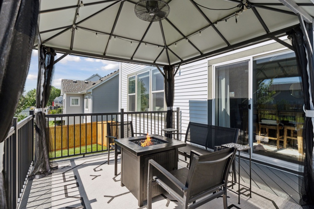 A covered patio on Fairchild Way with black metal railings, a fire pit table, four black chairs, and curtains. The patio overlooks a grassy backyard and neighboring houses under a blue sky—perfect for real estate photos.
