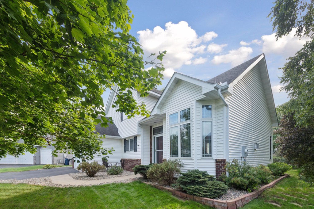 A white suburban house at 1648 Whitehall Place features large windows, dark shingles, and brick accents amid green lawns, shrubs, and trees under a partly cloudy sky. A curved walkway leads to the front door.