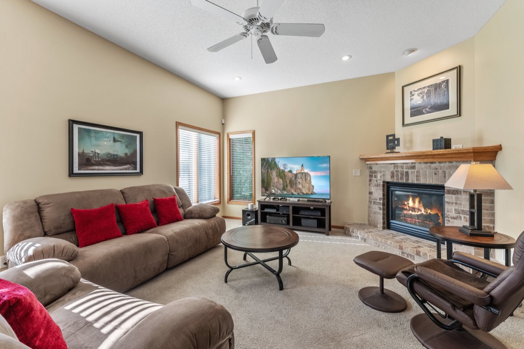 Cozy living room at 1648 Whitehall Place with beige walls, brown sofas with red pillows, a round coffee table, TV on a stand, brick fireplace with burning fire, artwork on walls, and large windows with blinds letting in natural light.