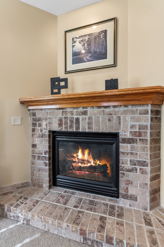 A brick fireplace with a burning fire, a wooden mantel above it, and decor including a framed landscape photo from PHOTOS Whitehall Place, all set against a beige wall.