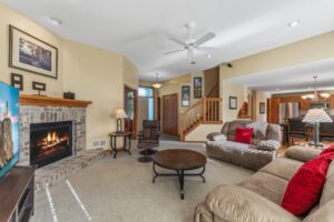 A cozy living room at Whitehall Place with a lit fireplace, red pillows on couches, a round coffee table, and a TV. The beige walls, carpeted floors, and open view to the kitchen highlight its real estate appeal. See PHOTOS 1648 for more details.