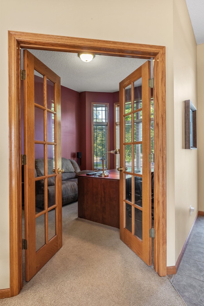 French doors with glass panes open to a home office at 1648 Whitehall Place, featuring a wooden desk, chair, brown sofa, and large windows with blinds. The room has red accent walls and beige carpeting; see photos for more details.