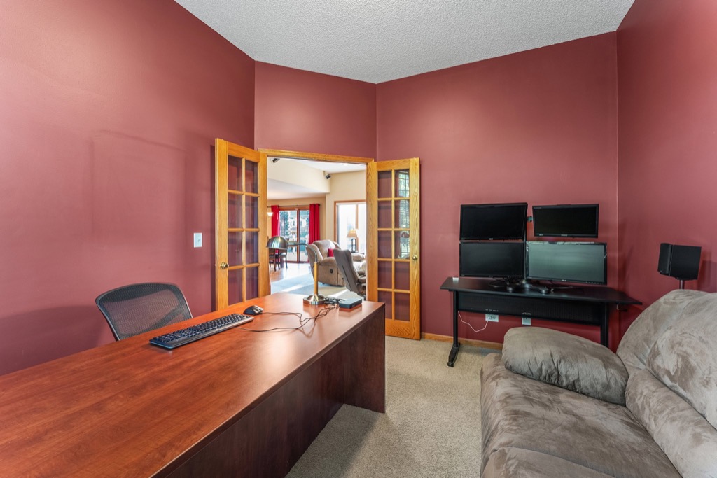 A home office with a wooden desk, office chair, computer monitors, and a couch. The walls are painted red, and double glass doors open to a bright adjoining room—perfect for displaying photos at 1648 Whitehall Place.