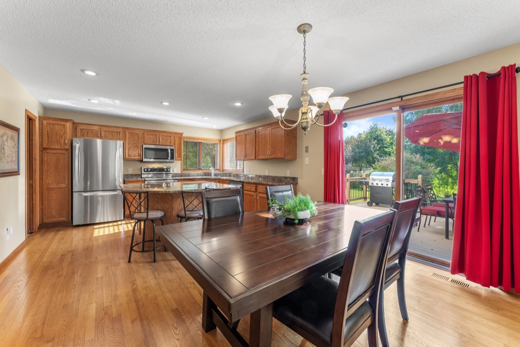 A kitchen and dining area at 1648 Whitehall Place features wood floors and cabinets, stainless steel appliances, a dark wood dining table with black chairs, a chandelier, and sliding glass doors with red curtains opening to a backyard deck with an umbrella.