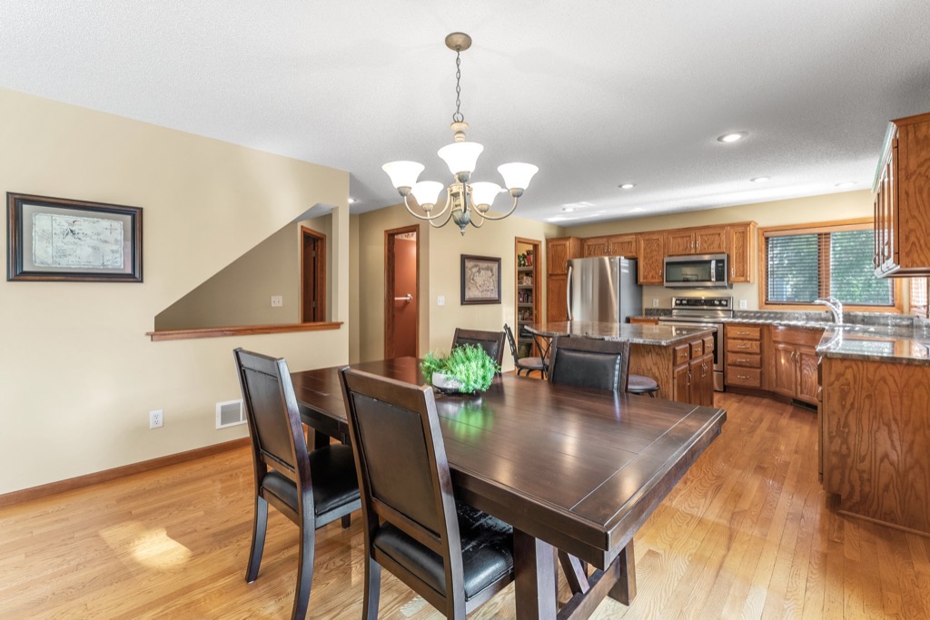 A spacious kitchen and dining area at 1648 Whitehall Place features wooden floors, a dark wooden table with six chairs, stainless steel appliances, wooden cabinets, a chandelier, and large windows letting in natural light. View PHOTOS for more details.