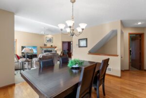 A dining room with a dark wooden table and chairs is adjacent to a living area featuring armchairs, a fireplace, a TV, and neutral-colored walls with wood accents and hardwood flooring—perfect for real estate at Whitehall Place. PHOTOS 1648.