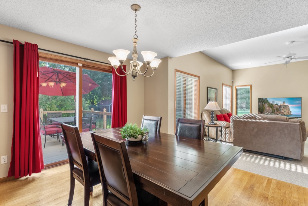 A dining area with a dark wooden table and chairs sits beside sliding glass doors with red curtains at Whitehall Place, opening to a deck with a large red umbrella. The living room and a TV are visible in the background. PHOTOS 1648.