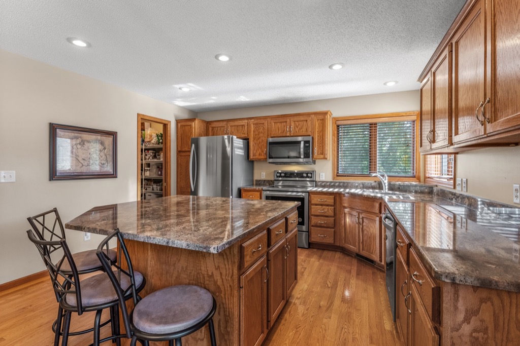 A spacious kitchen at Whitehall Place features wood cabinets, stainless steel appliances, dark marble countertops, a large island with two chairs, wood flooring, and a window above the sink letting in natural light. See photos for more details.