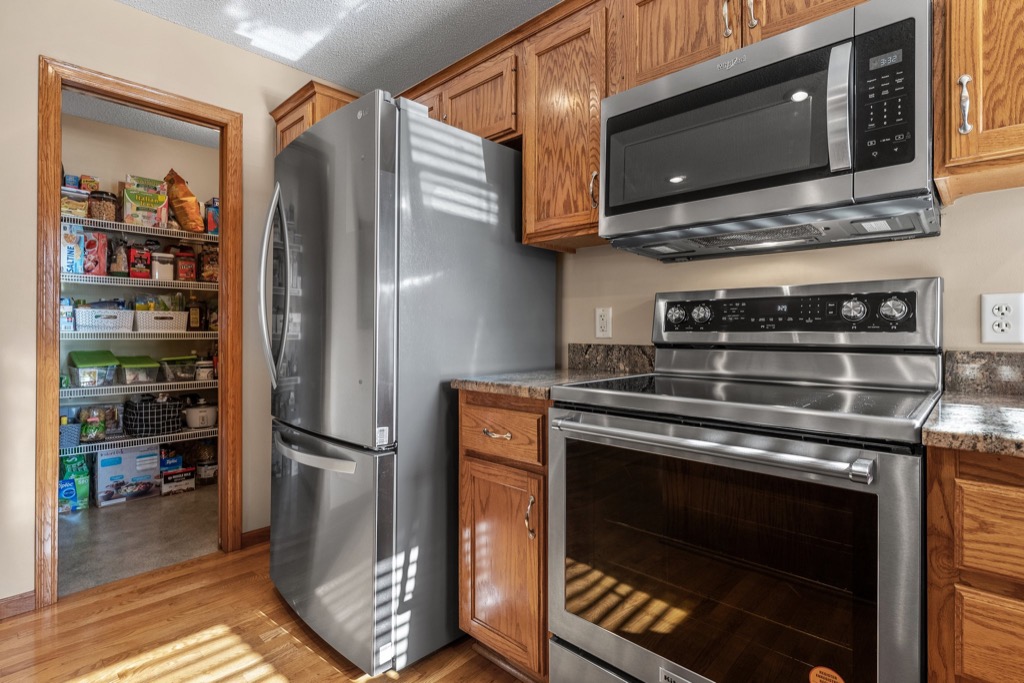 Modern kitchen at Whitehall Place featuring a stainless steel refrigerator, oven, and microwave, wooden cabinets, and a pantry stocked with food and storage containers. Sunlight casts shadows on the appliances and floor. See more in PHOTOS 1648.