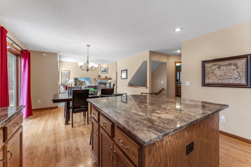 Spacious open-plan kitchen and dining area at 1648 Whitehall Place with a large island, wood cabinets, granite countertops, wood flooring, beige walls, and a dining table with chairs by large windows and red curtains. PHOTOS available.