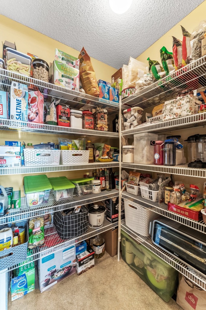 A well-organized pantry at 1648 Whitehall Place features white wire shelves filled with food items, snacks, boxed goods, containers, and baskets—all neatly arranged against yellow walls and a beige carpeted floor. See PHOTOS for details.