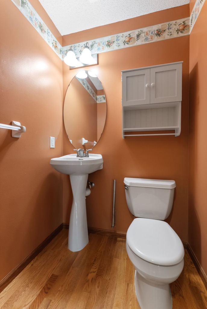 Charming small bathroom in a Whitehall Place property features orange walls, a pedestal sink beneath an oval mirror, white toilet, wood flooring, wall-mounted cabinet, floral wallpaper trim near the ceiling, and two lights above the mirror. See PHOTOS 1648.