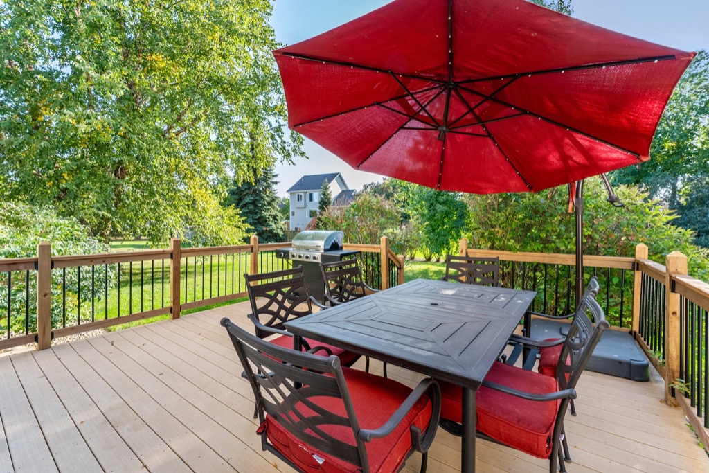 A wooden deck at 1648 Whitehall Place features black metal patio furniture with red cushions, a large red umbrella, and a grill, all overlooking a lush backyard with trees and a distant house on a sunny day.