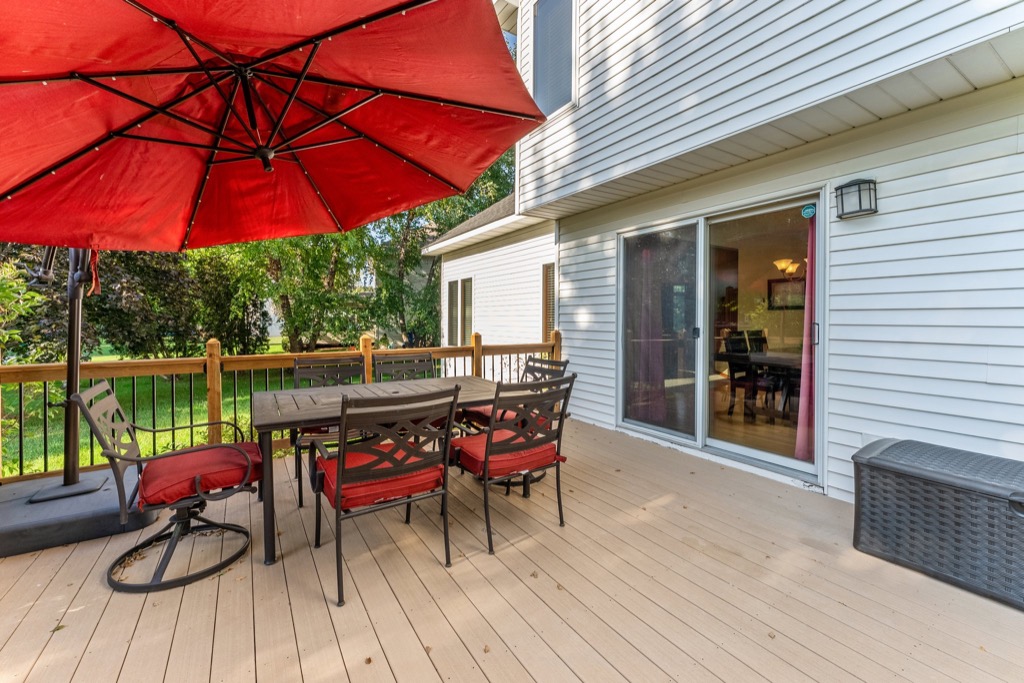 A wooden backyard deck at 1648 Whitehall Place features a dining table, six red-cushioned chairs, a large red umbrella, a black storage box, and a sliding glass door leading into the white-sided house, with green lawn and trees beyond.