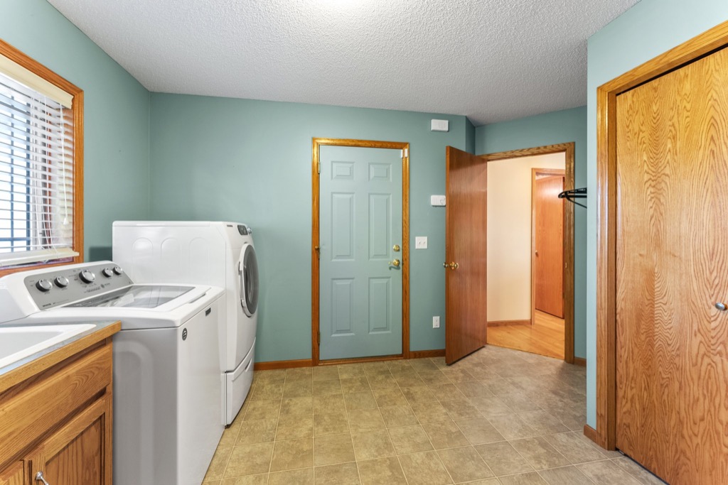 Laundry room at 1648 Whitehall Place with a washing machine and dryer, wood cabinets, greenish-blue walls, beige tile floor, wooden trim, a window with blinds, PHOTOS show two wooden doors—one open to a hallway.