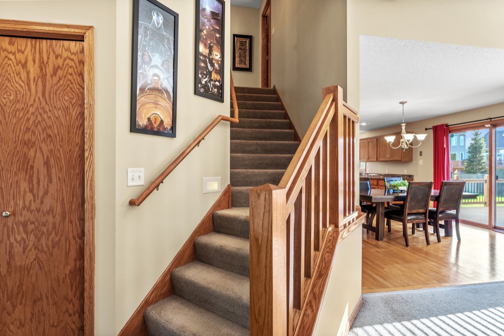 A carpeted staircase with a wooden railing leads up from a hallway adorned with framed art. To the right, an open dining area with a table, chairs, and large sliding glass doors overlooks the sunny backyard at 1648 Whitehall Place.