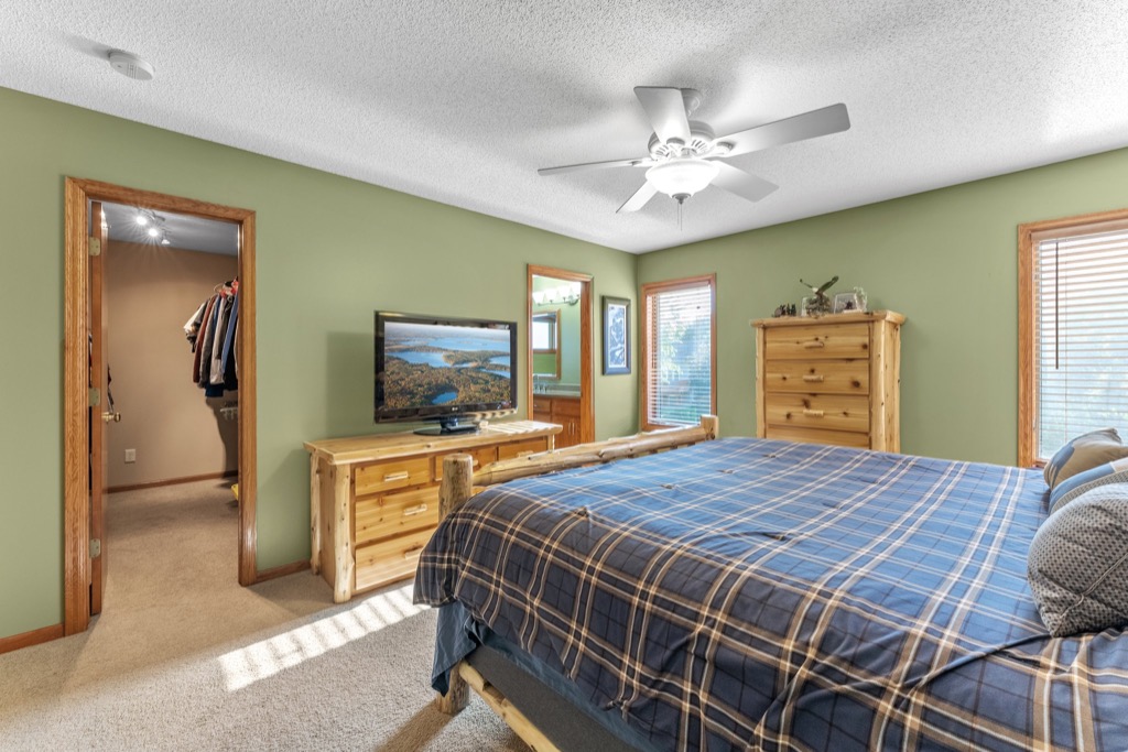 A cozy bedroom at 1648 Whitehall Place features green walls, a large bed with blue plaid bedding, wooden furniture, a ceiling fan, TV on a dresser, and two sunlit windows. An open door leads to the walk-in closet—see PHOTOS Whitehall Place for more.