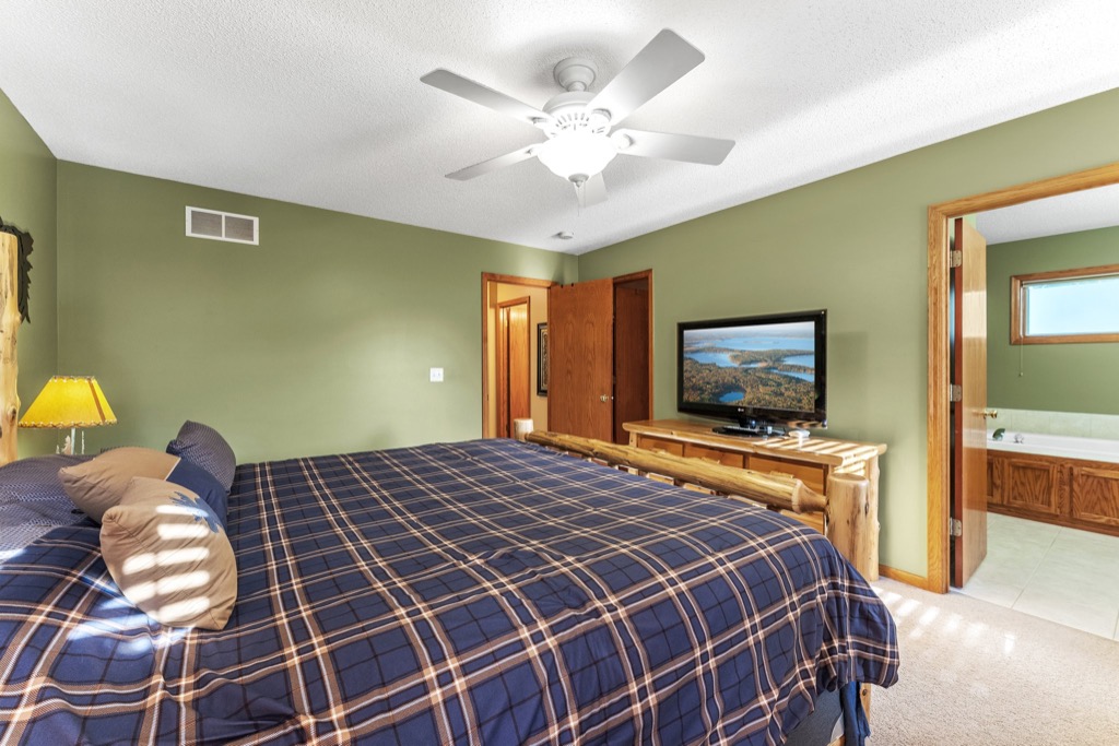A bedroom at Whitehall Place with green walls, a ceiling fan, a large bed with a plaid blue comforter, a wooden dresser with a TV, and an open door to a bathroom with a bathtub. See more in the Whitehall Place photos.
