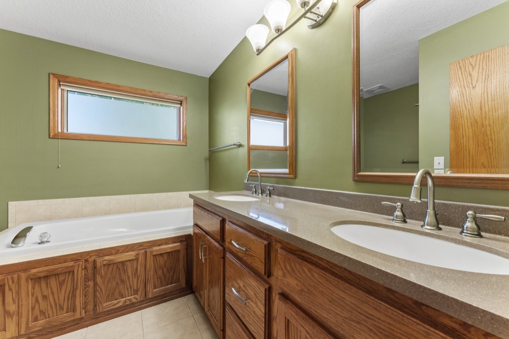 Spacious bathroom at Whitehall Place with olive green walls, wood trim, a large vanity with double sinks, two mirrors, overhead lights, and a bathtub with tiled surround beneath a frosted window. See PHOTOS 1648 for more real estate details.