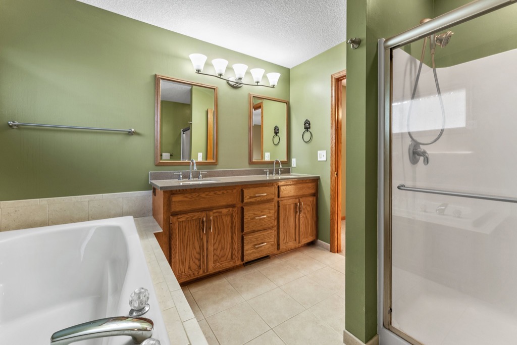 A bathroom at 1648 Whitehall Place features green walls, a double vanity with two mirrors and wooden cabinets, a bathtub, and a glass-enclosed shower with mounted showerhead. Beige tile flooring runs throughout the room.