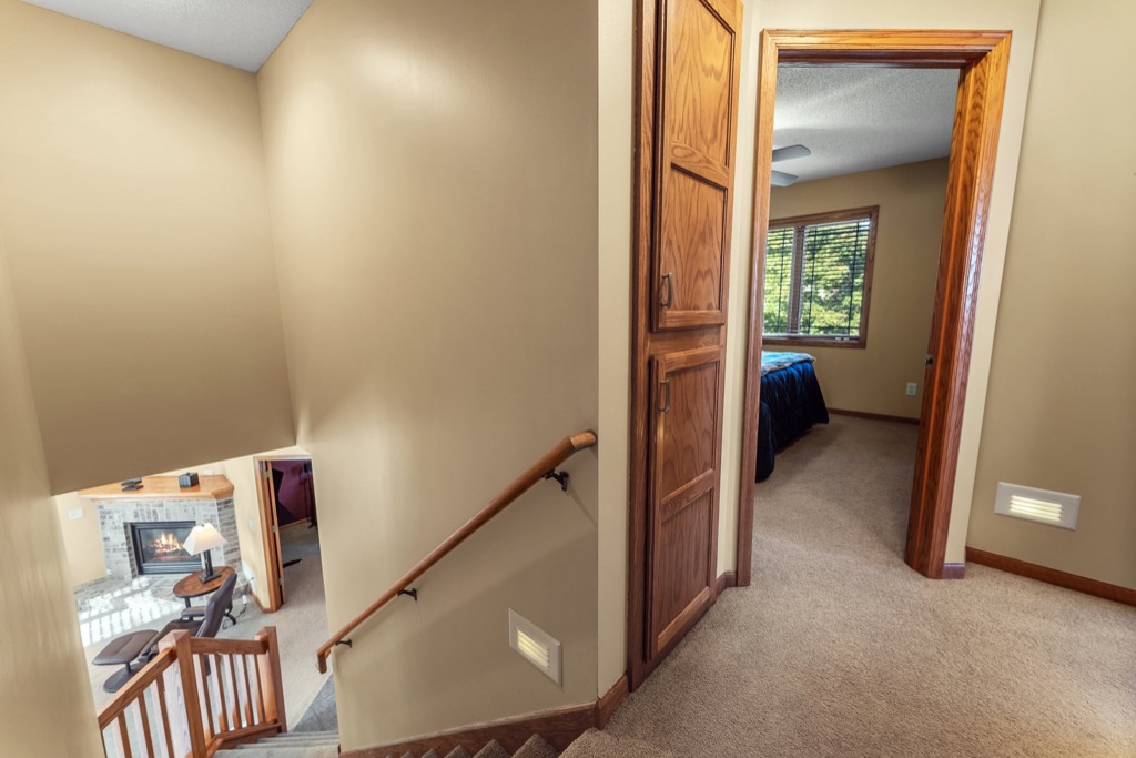 A stairway with a wooden handrail leads upstairs to a hallway with beige walls and carpet at 1648 Whitehall Place. An open door reveals a bedroom, while downstairs, a fireplace and seating area complete the inviting atmosphere.