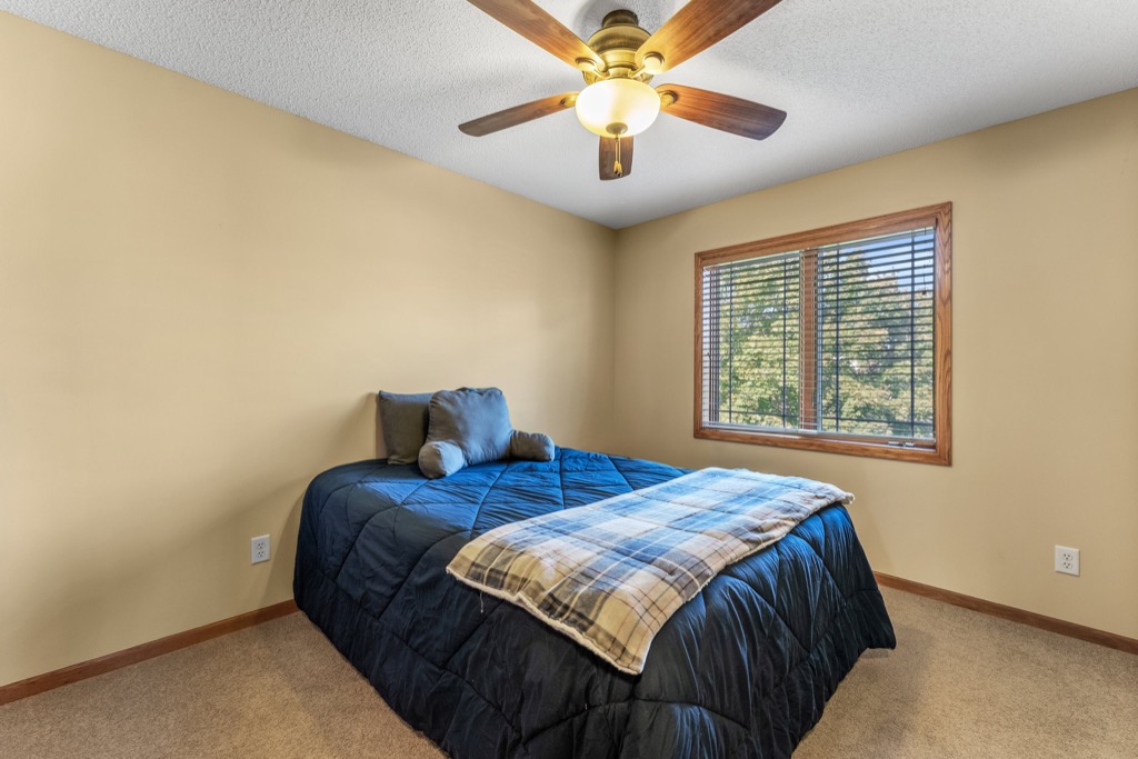 A simple bedroom at 1648 Whitehall Place with beige walls, a ceiling fan, a window with blinds, and a bed covered in a dark blue comforter and plaid blanket, topped with two decorative pillows against the wall. See PHOTOS for details.