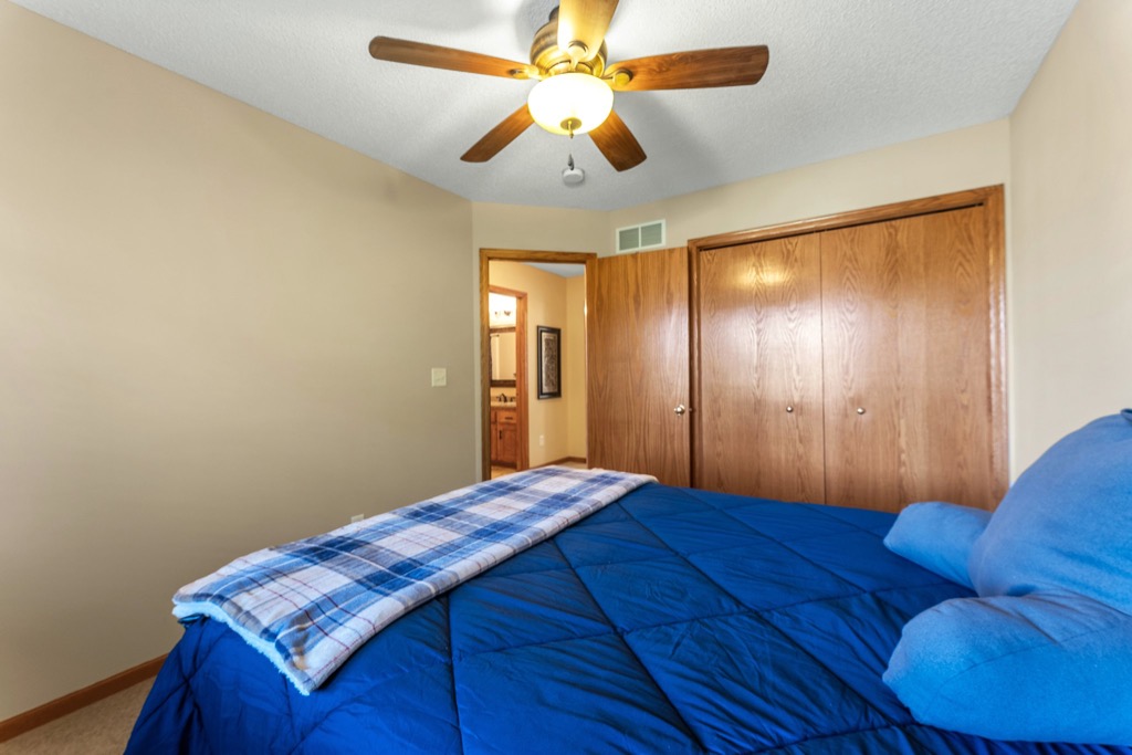 A bedroom at Whitehall Place, 1648, with beige walls features a bed with a blue comforter and plaid blanket, wooden closet doors, a ceiling fan with a light, and an open door to a bathroom—perfect for cozy photos.