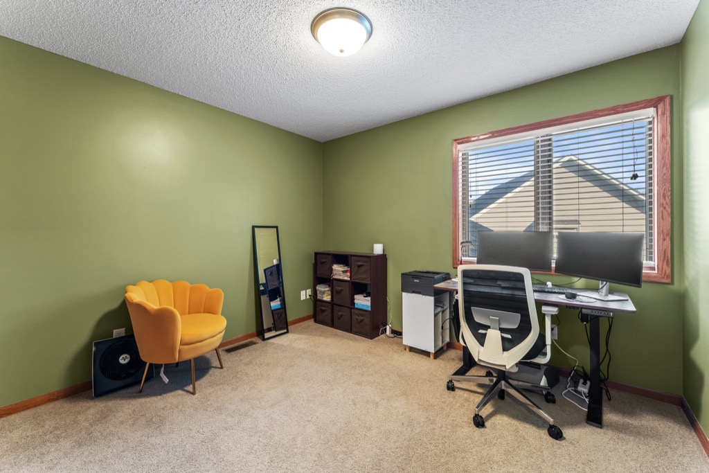 A home office at 1648 Whitehall Place features green walls, beige carpet, a yellow accent chair, a standing mirror, small shelving unit, dual-monitor desk with white office chair, and a window streaming natural light—see more in the property photos.