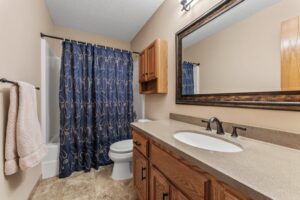 A bathroom at 1648 Whitehall Place features a wooden vanity, beige countertop, and double faucet sink. A large framed mirror hangs above. The shower has a blue patterned curtain, and two beige towels hang on a rack beside the tub. PHOTOS available.