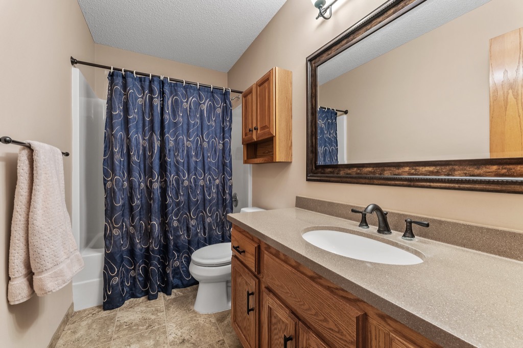A bathroom at 1648 Whitehall Place features a wooden vanity, beige countertop, and double faucet sink. A large framed mirror hangs above. The shower has a blue patterned curtain, and two beige towels hang on a rack beside the tub. PHOTOS available.