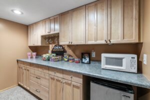 A home snack bar at Whitehall Place features light wood cabinets, a gray countertop, a microwave, mini fridge, popcorn containers, candy, snacks, framed PHOTOS 1648, and hanging wine glasses against tan walls—perfect for real estate showings.