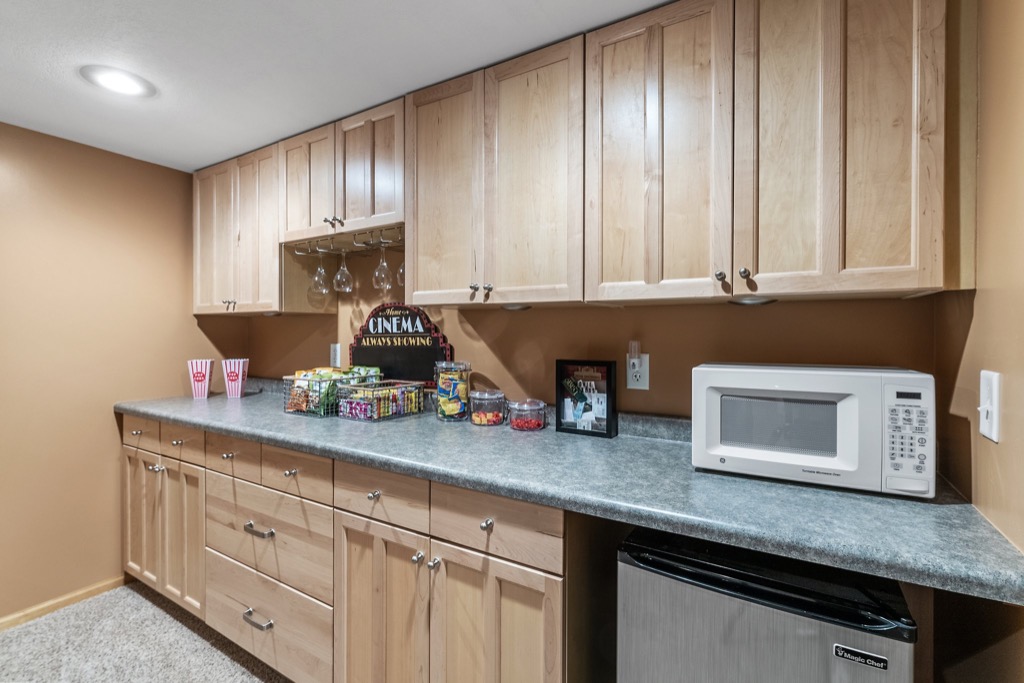 A home snack bar at Whitehall Place features light wood cabinets, a gray countertop, a microwave, mini fridge, popcorn containers, candy, snacks, framed PHOTOS 1648, and hanging wine glasses against tan walls—perfect for real estate showings.