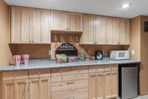 A home snack bar at Whitehall Place with light wood cabinets, a gray countertop, microwave, mini fridge, popcorn tubs, movie-style snacks, and a “Cinema Always Showing” sign under hanging wine glasses. PHOTOS 1648.