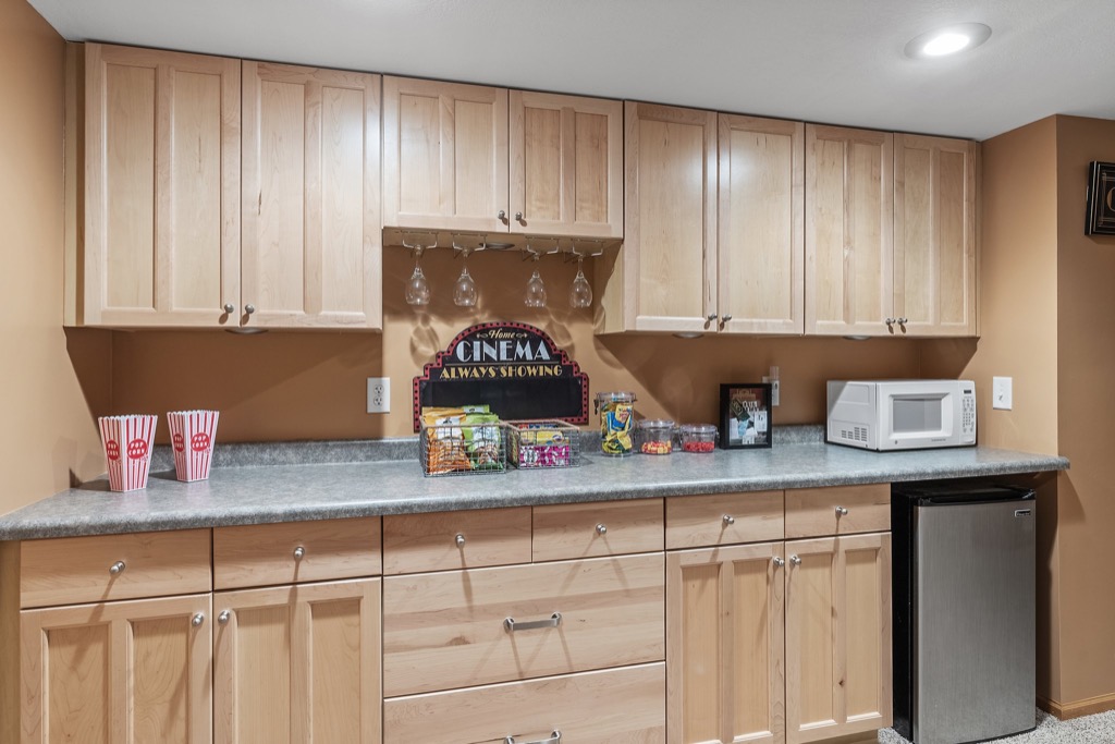 A home snack bar at Whitehall Place with light wood cabinets, a gray countertop, microwave, mini fridge, popcorn tubs, movie-style snacks, and a “Cinema Always Showing” sign under hanging wine glasses. PHOTOS 1648.