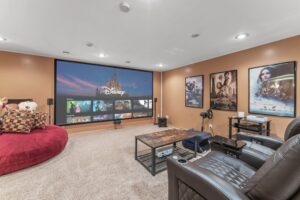 A cozy home theater room at 1648 Whitehall Place features a large screen displaying Disney+ content, brown walls, movie posters, leather recliners, a round red sofa with plush toys, a wooden coffee table, and carpeted floors. PHOTOS available.