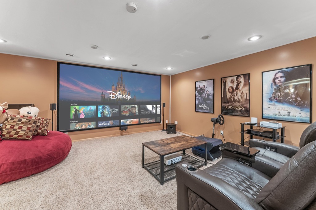 A cozy home theater room at 1648 Whitehall Place features a large screen displaying Disney+ content, brown walls, movie posters, leather recliners, a round red sofa with plush toys, a wooden coffee table, and carpeted floors. PHOTOS available.