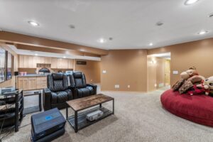 A cozy basement room at 1648 Whitehall Place with tan walls and carpet, featuring two black recliners, a large red bean bag with teddy bears, a wooden coffee table, and a kitchenette. Bright ceiling lights illuminate the space—see PHOTOS for details.