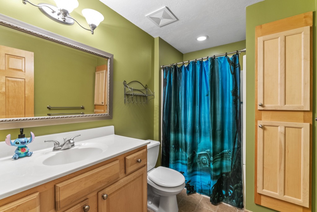 A bathroom at 1648 Whitehall Place features olive green walls, wooden cabinets, a white sink, a toilet, and a blue shower curtain with a design. A small Stitch figurine sits on the countertop near the faucet.
