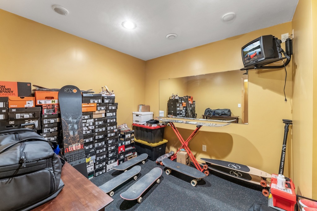 A room with yellow walls at 1648 Whitehall Place contains several skateboards, a snowboard, stacks of shoe boxes, storage bins, a folding table, a wall mirror, and PHOTOS displayed next to a small TV mounted high in the corner.