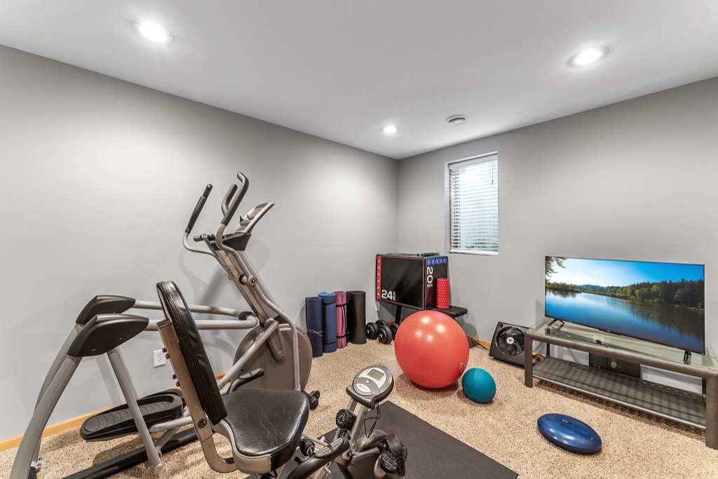 A home gym at Whitehall Place with an exercise bike, elliptical machine, stability balls, foam rollers, and mats. The space features a TV on a stand, window, beige carpet, light gray walls, and recessed ceiling lights—ideal for real estate PHOTOS 1648.