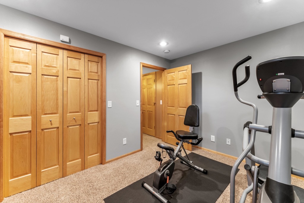 A home gym room at 1648 Whitehall Place features beige carpet, gray walls, wooden double doors, an exercise bike, and elliptical machine on black mats under ceiling lights. See more in PHOTOS Whitehall Place.