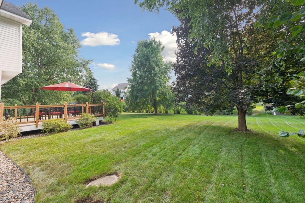 A backyard at Whitehall Place with a well-maintained green lawn, several large trees, and a wooden deck on the left side featuring a red patio umbrella and outdoor seating. The blue sky with scattered clouds enhances this PHOTOS 1648 scene.