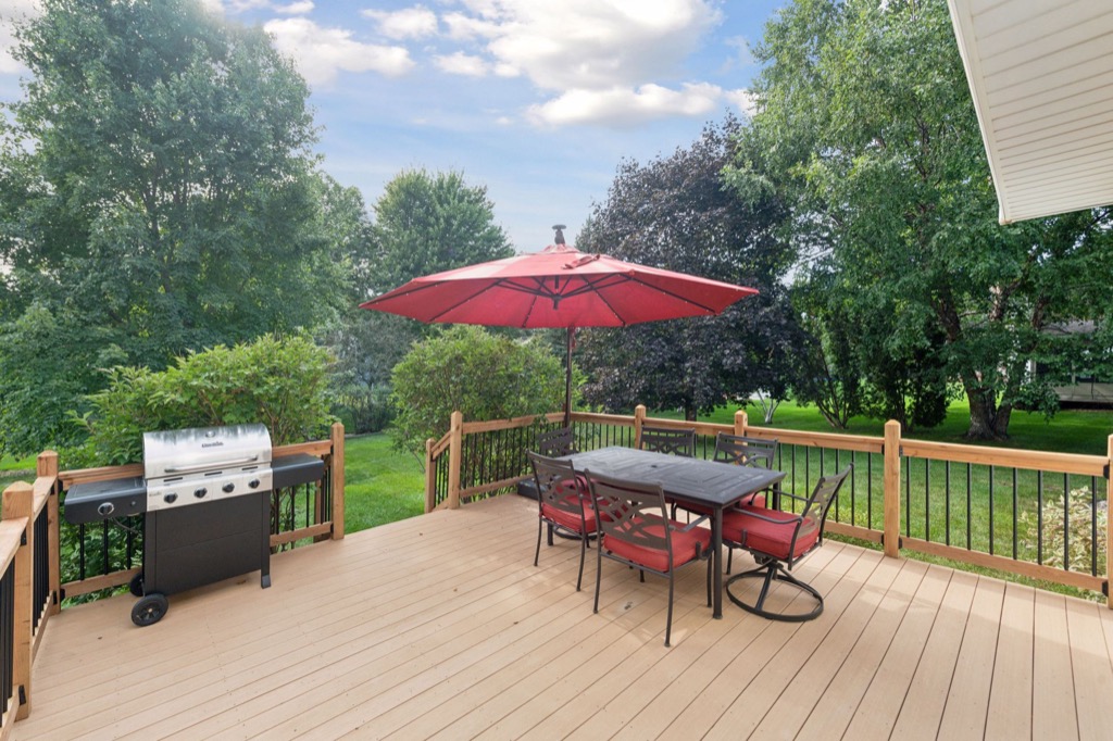 A backyard deck at 1648 Whitehall Place features a dining table, red chairs, and a large red umbrella beside a gas grill. The deck overlooks a green lawn with trees and bushes under a partly cloudy sky—see PHOTOS for more.