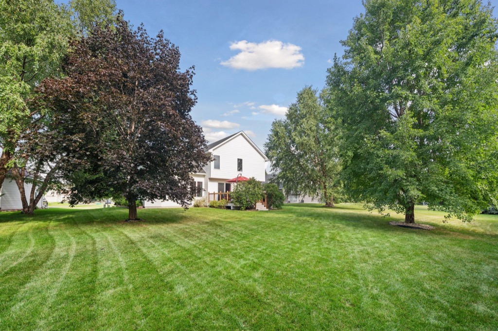 A white house, reminiscent of Whitehall Place, with a porch and red umbrella sits in the background, surrounded by large green and dark-leaved trees on a manicured lawn under a blue sky with a few clouds—perfect for memorable PHOTOS.
