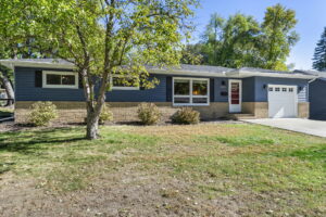 A single-story house with blue siding, tan brick accents, a red front door, white-framed windows, attached garage, and well-kept lawn with a tree and shrubs in the front yard on a sunny day.