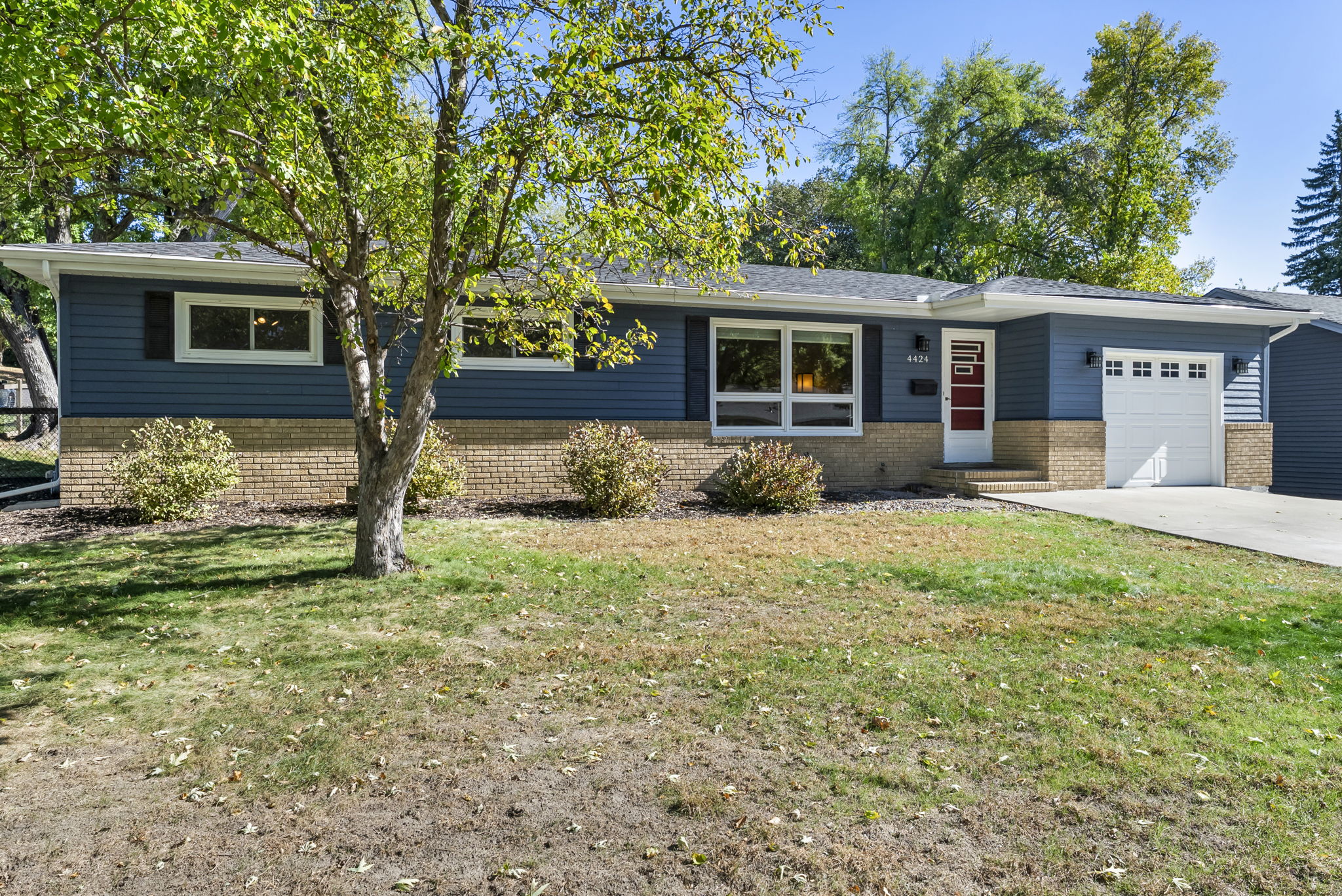 A single-story house with blue siding, tan brick accents, a red front door, white-framed windows, attached garage, and well-kept lawn with a tree and shrubs in the front yard on a sunny day.