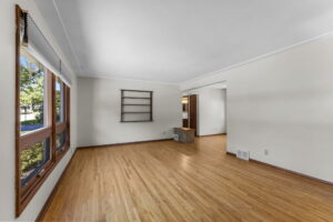 A spacious, empty living room with light wood floors, large front windows, white walls, a small built-in wall shelf, and an open doorway leading to another room. Natural light fills the space.