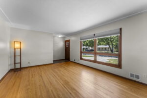 Bright, empty living room with light wood floors, beige walls, a large window showing trees and houses outside, a modern floor lamp in the corner, and a wooden front door with three vertical windows.