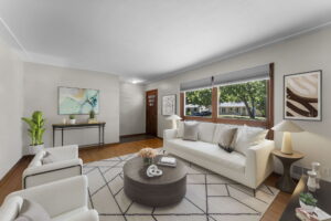 A modern living room with a white sofa, two armchairs, a round coffee table on a patterned rug, and large windows showing trees outside. There is wall art, a console table, and potted plants decorating the space.